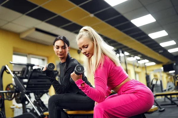 Тренер-женщина показывает клиенту упражнения для бицепсов с гантелями, сидя на скамейке в тренажерном зале — стоковое фото
