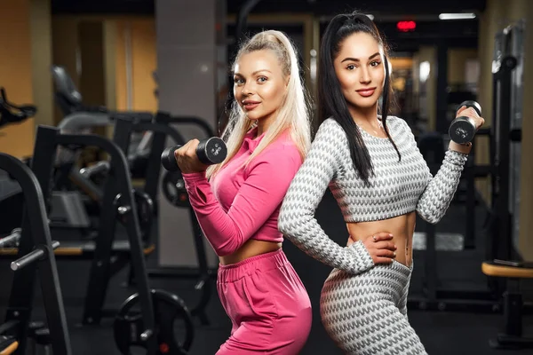 Twee sportmeisjes doen oefeningen voor biceps met halters terwijl ze staan en poseren in de sportschool. Fitness — Stockfoto
