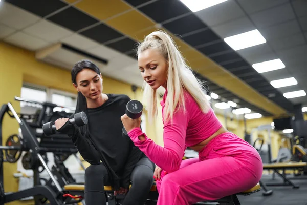 Kobieta trener pokazuje dziewczynie ćwiczenie na bicepsy z hantlem podczas siedzenia na ławce w siłowni — Zdjęcie stockowe