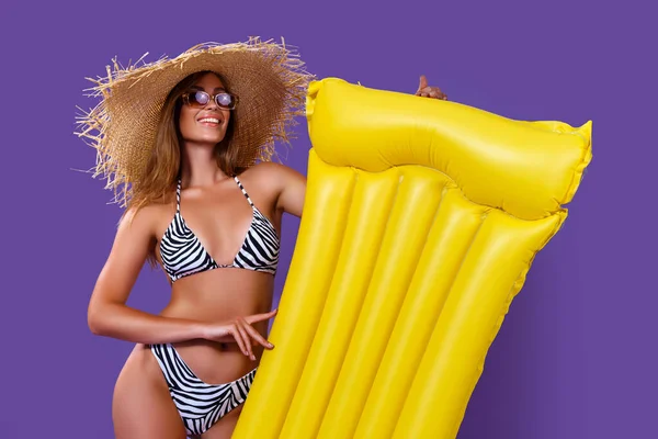 Cheerful girl in bikini going to poll with yellow inflatable mattress, purple studio background — Stock Photo, Image