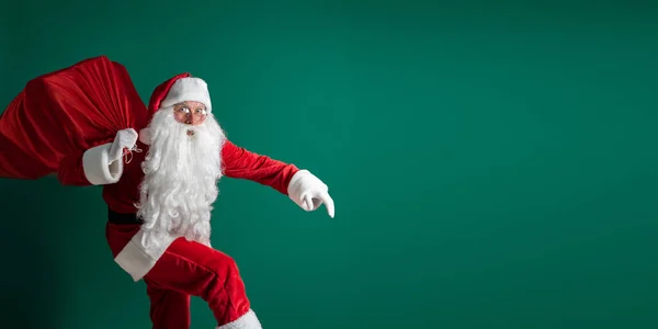 Sorprendido Santa Claus a hurtadillas mientras llevaba un enorme saco rojo con regalos sobre fondo de estudio verde —  Fotos de Stock