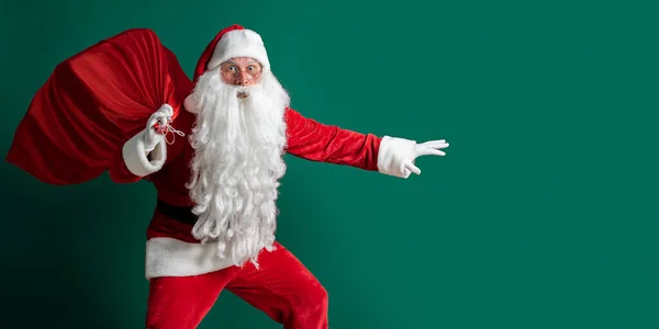 Emocjonalne Santa Claus skradanie się niosąc ogromny czerwony worek z prezentami na zielonym tle studio — Zdjęcie stockowe