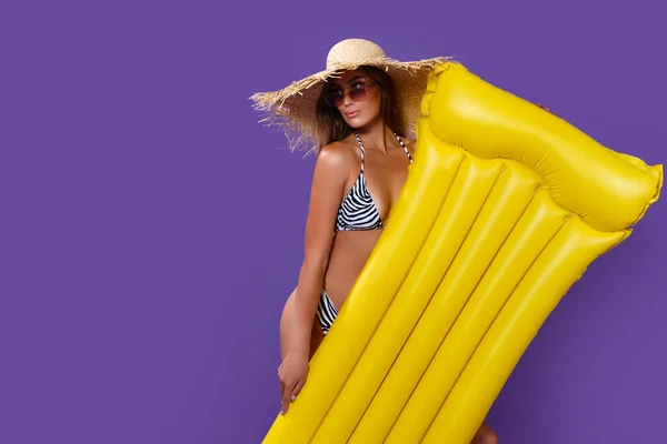 Atrakcyjna dziewczyna w bikini w słomkowym kapeluszu trzyma żółty materac pływacki stoi na fioletowym tle — Zdjęcie stockowe