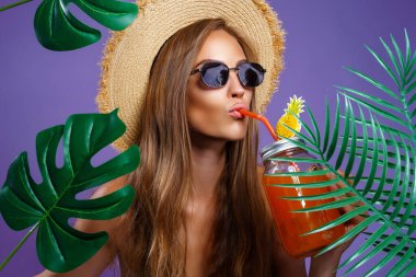 Yaz aylarında meyve kokteyli içen, son moda güneş gözlüğü takan, hasır şapka takan ve mor stüdyo arka planında izole tropik bitkilerin yanında duran güzel bir kadının moda portresi. 