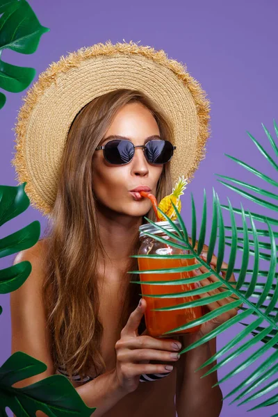 Attraktives Mädchen trinkt Fruchtcocktail in Sonnenbrille und Strohhut, während sie neben tropischen Pflanzen steht — Stockfoto