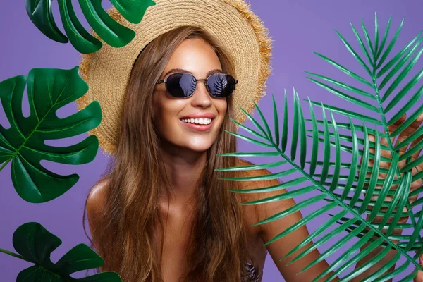 Портрет усміхненої дівчини носить солом'яний капелюх бікіні та сонцезахисні окуляри, які стоять між тропічними рослинами — стокове фото