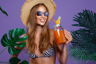 Mutlu güzel kız elinde güneş gözlüğü ve hasır şapkayla meyve kokteyli tutarken tropik bitkilerin yanında duruyor.