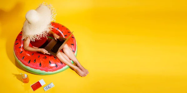 Gadis bertopi jerami duduk di inflatable watermelon ring menggunakan pc pada latar belakang kuning. bepergian ke luar negeri — Stok Foto