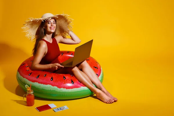Happy girl dengan topi jerami duduk di inflatable watermelon ring menggunakan pc pada latar belakang kuning. penjelajah. — Stok Foto