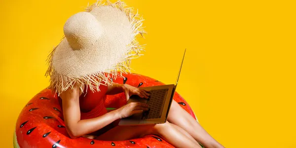 Wanita bertopi jerami duduk di cincin tiup bekerja pada laptop dengan latar belakang kuning. Pekerja lepas. Perjalanan — Stok Foto