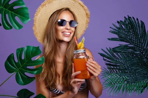 Усміхнена дівчина тримає фруктовий коктейль в сонцезахисних окулярах і солом'яному капелюсі, поки стоїть біля тропічних рослин — стокове фото