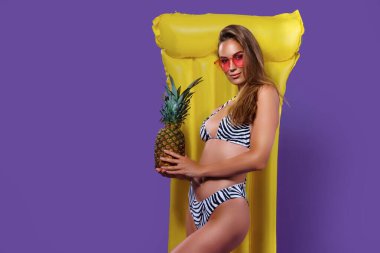 Güzel beyaz kadın ananas tutarken bikinili, sarı şişme yataklı poz veriyor.. 