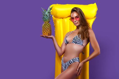 Genç beyaz kadın ananas tutarken bikinili, sarı şişme yataklı poz veriyor.. 