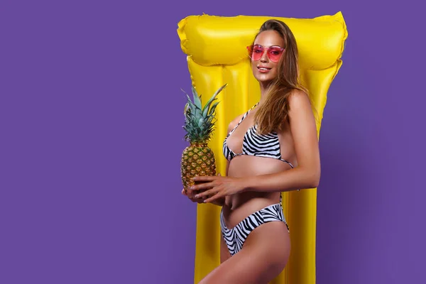 Young slim caucasian girl posing in bikini with yellow inflatable mattress while holding pineapple. — Stock Photo, Image