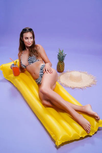Smiling woman wears in bikini lyes on inflatable matress while holds cocktail on purple background — Stock Photo, Image
