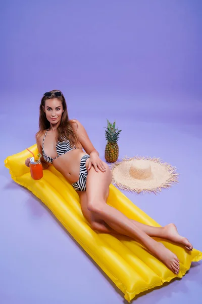Happy woman wears in bikini lyes on inflatable matress while holds cocktail on purple background — Stock Photo, Image