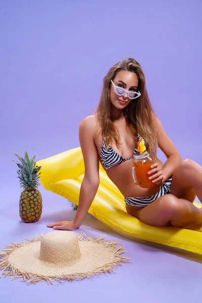 Smiling woman wears in bikini sits on inflatable matress whith fruit cocktail on purple background — Stock Photo, Image