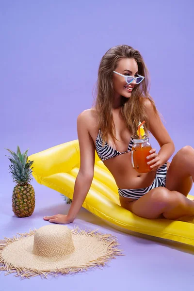 Fröhliche Frau im Bikini sitzt auf aufblasbarer Matratze mit Fruchtcocktail auf lila Hintergrund — Stockfoto