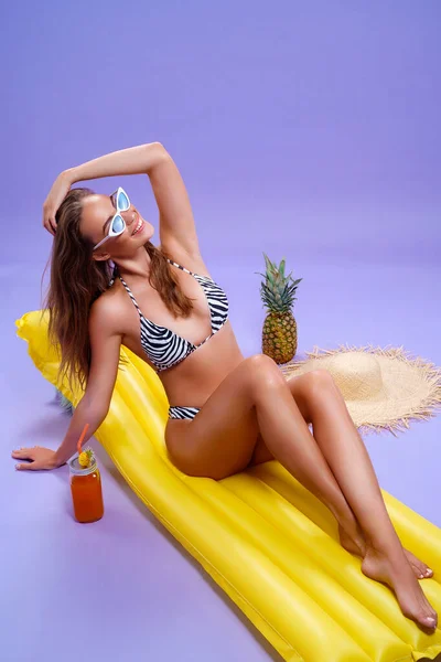 Bikinili harika bir kadın mor arka planda şişme matress beyaz meyve kokteyli üzerine oturur. — Stok fotoğraf