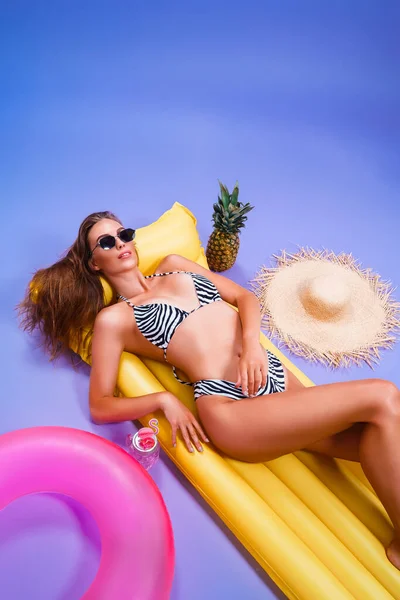 Sexy kobieta nosi w bikini w okularach przeciwsłonecznych leżących na żółty dmuchany materac na fioletowym tle — Zdjęcie stockowe