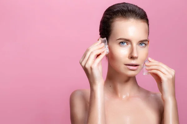 Mooie brunette jonge naakte vrouw met perfecte huid houdt ijsblokjes in de buurt gezicht op roze achtergrond — Stockfoto