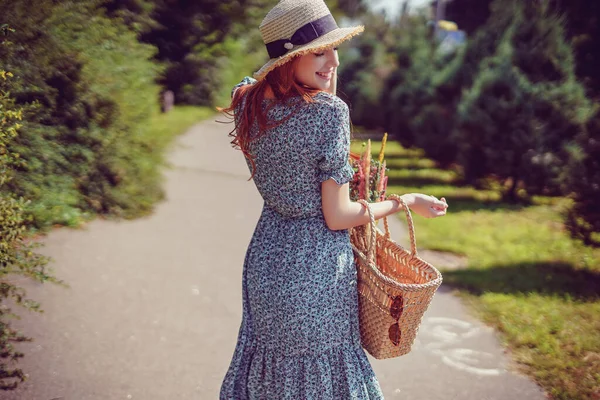 Back view of adult redhead girl with stylish straw bag and straw hat who walks in park outdoor — Stock Photo, Image