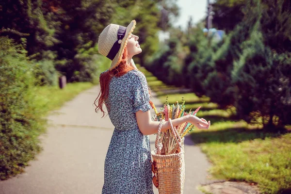 Vue arrière de la jeune rousse femme avec sac de paille élégant et chapeau de paille qui marche dans le parc en plein air — Photo