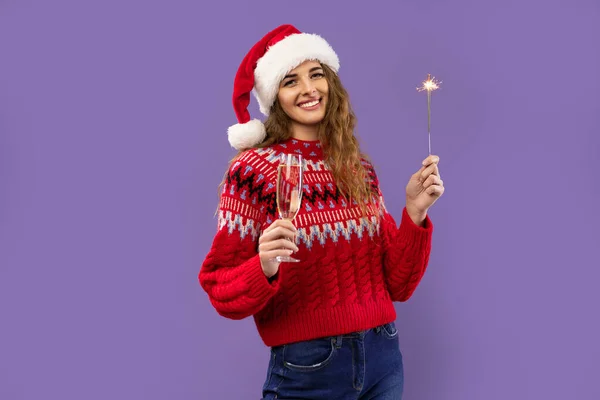 Smiling Santa girl holds glass of champagne and sparkler on purple background. Christmas party — Stock Photo, Image