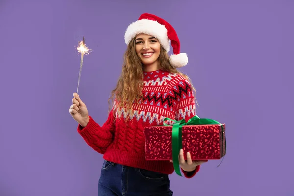 Happy young woman wears Santa hat holds gift box and sparkler on purple background. Christmas party — Stock Photo, Image