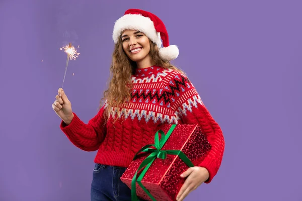 Happy beauty girl wears Santa hat holds gift box and sparkler on purple background. Christmas party — Stock Photo, Image