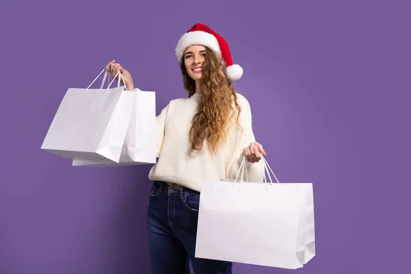 Glad ung kvinna bär Santa hatt håller shoppingväskor medan ler mot kameran på violett bakgrund — Stockfoto