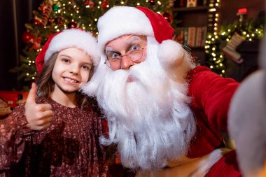 Noel Baba, baş parmağını gösteren sevimli küçük bir kızla selfie çekiyor. Noel arkaplanı