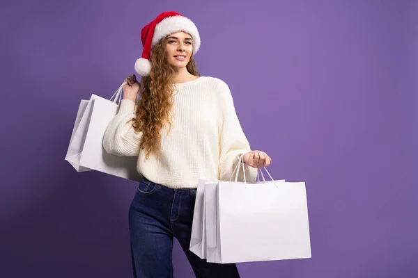 Smiling young woman wears Santa hat holds shopping bags while smiling at camera on purple background — Stock Photo, Image
