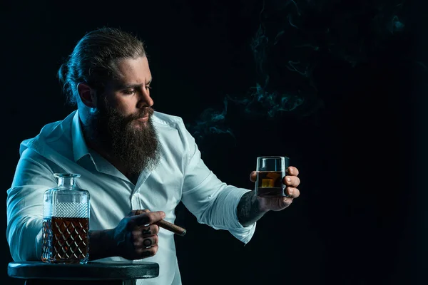 Przystojny brodaty samiec pali cygaro trzyma szklankę whisky, ubraną w białą koszulę. Zdjęcia studyjne — Zdjęcie stockowe