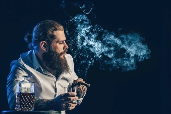 Przystojny brodaty facet pali cygaro trzyma szklankę whisky, dmucha dymem. Zdjęcia studyjne — Zdjęcie stockowe