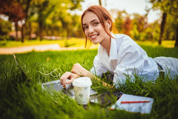 Красива усміхнена студентка використовує ноутбук, лежачи на траві на відкритому повітрі. Концепція освіти — стокове фото