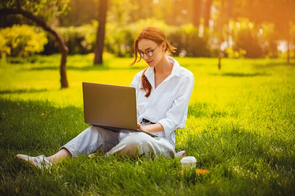 Рудий фрілансер дівчата окуляри працює з ноутбуком, на відкритому повітрі, сидячи на траві. Фріланс — стокове фото