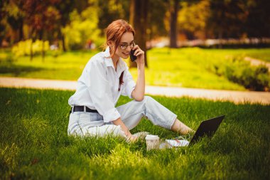 Parktaki çimlerin üzerinde dizüstü bilgisayarıyla oturan ve cep telefonuyla konuşan mutlu genç kız.