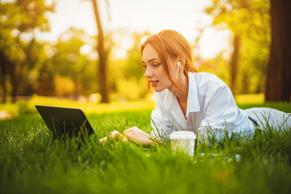 Молода руда студентка лежить на траві, використовує ноутбук і навушники. Дистанційне навчання . — стокове фото