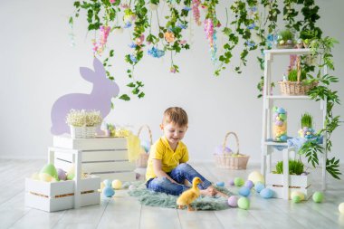 Gülen çocuk elinde küçük bir ördek yavrusuyla ahşap zeminde oturuyor. Çocuklar için Paskalya