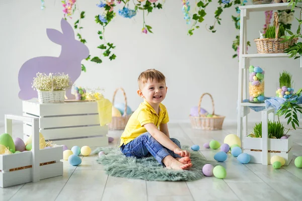 Cheerful Little boy sits on floor in Easter studio decoration with flowers, wooden rabbits, eggs — Stock Photo, Image
