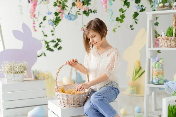 Cute girl communicates with ducklings in basket while sits on the wooden box. Easter decoration — Stock Photo, Image