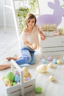 Beyaz kız Paskalya stüdyosunda yerde oturuyor. Çiçekler, ahşap tavşanlar, renkli yumurtalar.