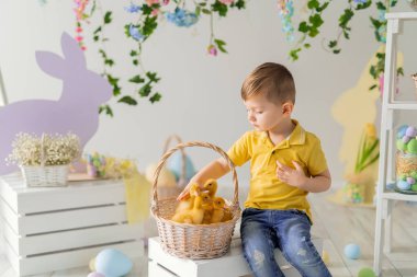 Küçük çocuk tahta kutunun üzerinde otururken ördek yavrularıyla sepetin içinde iletişim kuruyor. Paskalya dekorasyonu