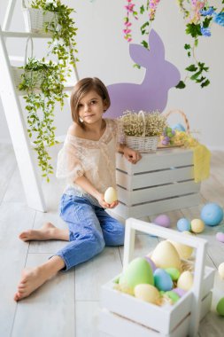 Beyaz kız Paskalya stüdyosunda yerde oturuyor. Çiçekler, ahşap tavşanlar, boyalı yumurtalar.