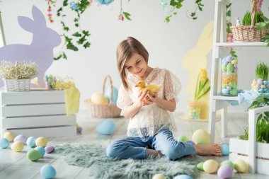 Paskalya kompozisyonu. Küçük kız ahşap zeminde otururken ördek yavrusuyla oynuyor ve iletişim kuruyor.