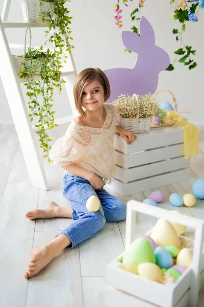 Cute girl sits on floor in Easter studio decoration with flowers, wooden rabbits, colorful dyed eggs — Stock Photo, Image