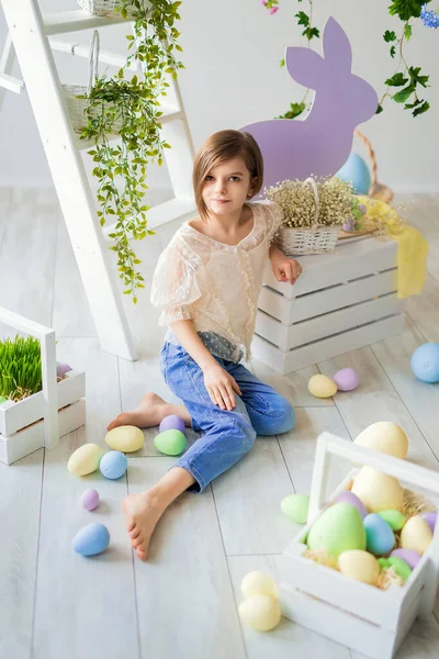 Küçük kız Paskalya 'da yerde oturuyor. Çiçekler, ahşap tavşan, renkli yumurtalar.