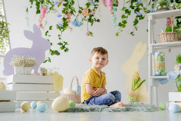 Μικρό αγόρι κάθεται σε ξύλινο πάτωμα στο στούντιο διακόσμηση Πάσχα με λουλούδια, ξύλινα κουνέλια, βαμμένα αυγά — Φωτογραφία Αρχείου