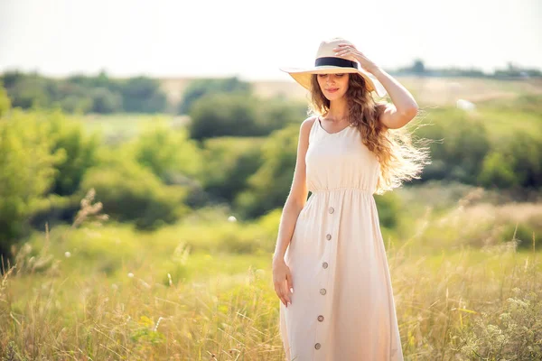 Feliz hermosa chica se para en el campo en verano mientras usa un sombrero de sol y vestido midi. Estilo de vida —  Fotos de Stock